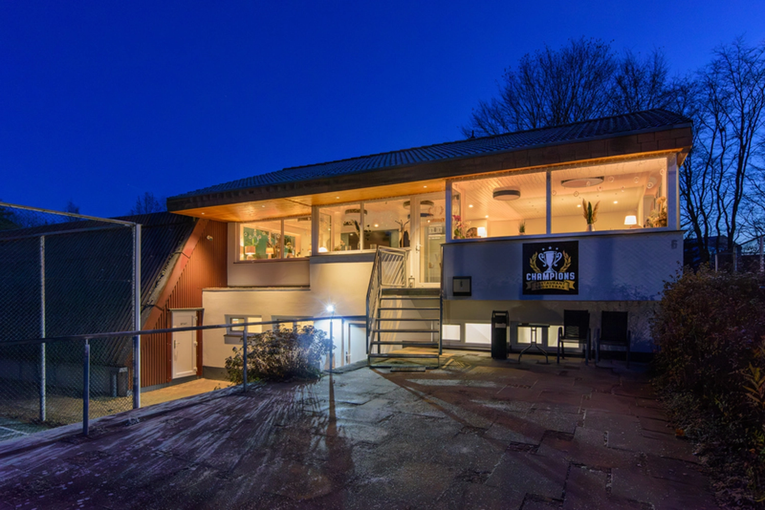 Champions Restaurant und Sportsbar - Genießen Sie frisch zubereitete Speisen, kühle Getränke - Ein modernes, mehrstöckiges Gebäude mit großen Fenstern und nachts beleuchtetem Innenraum, mit einer Außenterrasse und einem „CHAMPIONS“-Schild.