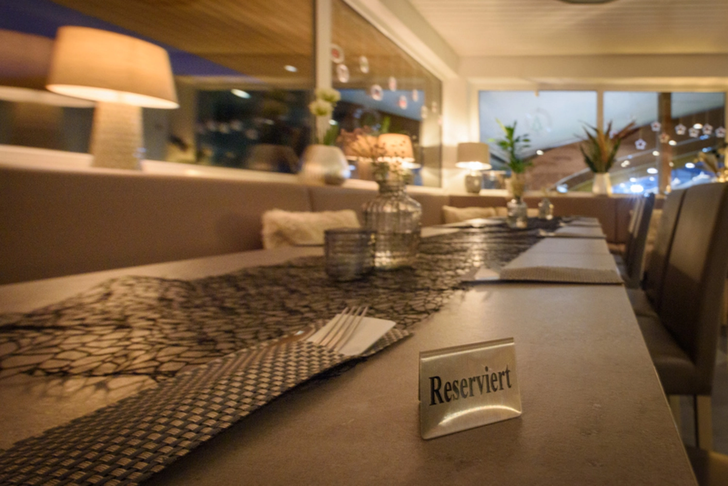 Champions Restaurant und Sportsbar - Genießen Sie frisch zubereitete Speisen, kühle Getränke - Nahaufnahme eines Restauranttisches mit einem silbernen „Reserviert“-Schild. Der Tisch ist gedeckt mit Platzdeckchen, Besteck, Gläsern und einer Wasserkaraffe; im Hintergrund ist das Restaurantinterieur verschwommen zu sehen.