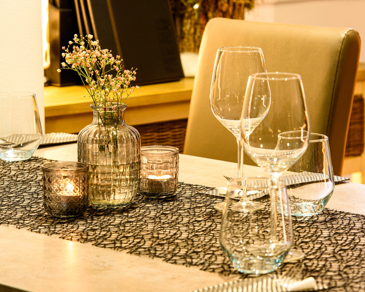 Champions Restaurant und Sportsbar - Genießen Sie frisch zubereitete Speisen, kühle Getränke - Eine Nahaufnahme eines gedeckten Esstisches mit mehreren Weingläsern, einer durchsichtigen Vase mit kleinen rosa und weißen Blüten und mehreren Teelichtern, alles auf einem dunklen, strukturierten Platzdeckchen oder Tischläufer. Die Beleuchtung ist warm und stimmungsvoll.