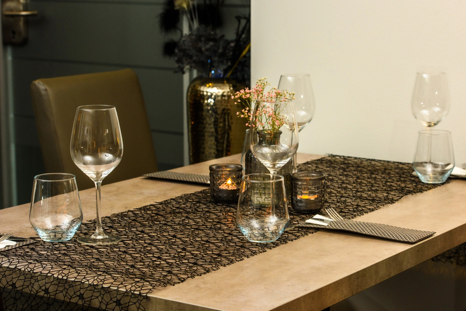 Champions Restaurant und Sportsbar - Genießen Sie frisch zubereitete Speisen, kühle Getränke - Eine Nahaufnahme eines gedeckten Esstisches mit mehreren Weingläsern, einer durchsichtigen Vase mit kleinen rosa und weißen Blüten und mehreren Teelichtern, alles auf einem dunklen, strukturierten Platzdeckchen oder Tischläufer. Die Beleuchtung ist warm und stimmungsvoll.